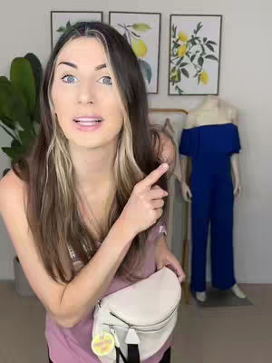 A woman with long brown hair, blonde highlights, and blue eyes points to her right. She wears a pink tank top and a white fanny pack. In the background, there are framed pictures and a mannequin wearing a blue off-the-shoulder jumpsuit.