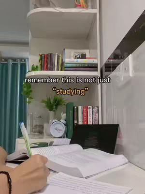 A person's hand writing on a book at a desk. In the background, there's a full white open shelf filled with books and some green fake plants. On the desk there is a black laptop, a white clock, and a pen.