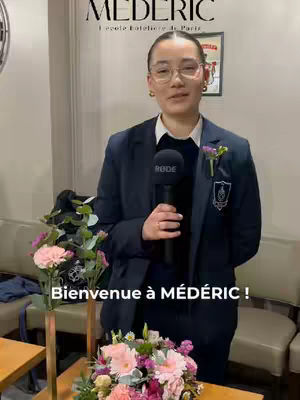 A woman wearing glasses and a blazer stands holding a microphone and surrounded by floral arrangements in front of a backdrop.