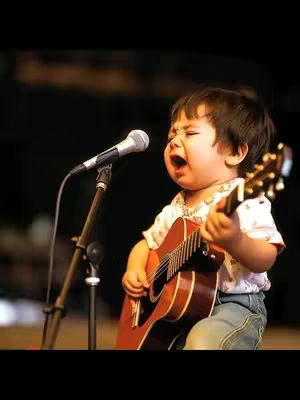 A young child, likely a toddler, is on a stage holding a guitar and singing into a microphone. The child is wearing a white collared shirt with a print and blue jeans, and is mid-song with their mouth open. A microphone stand is positioned to the side. The lighting is warm, creating a spotlight effect on the child. The background is a dark, blurred setting.
