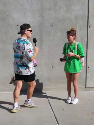A man in a floral shirt and sunglasses holding a microphone, interviewing a woman in a green shirt near a concrete wall. The man is positioned left, the woman is on the right with a backpack.