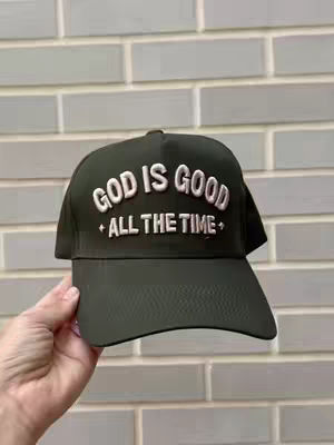 A close-up shot of a black baseball cap held by a hand. The cap has the phrase "GOD IS GOOD ALL THE TIME" embroidered in white, with the words arranged in a semi-circular design. The background is a light grey brick wall.