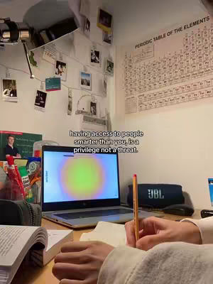 A well-lit room shows a laptop open on a wooden desk, with a person's hands visible in the foreground. Text overlaid in a white box highlights a motivational message. Adjacent to the laptop is a book and a pen, and a JBL speaker. In the background are photos and a periodic table chart.