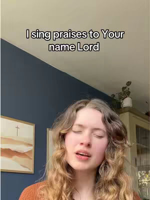 The cover image features a young woman with long, wavy blonde hair, her eyes closed, singing. The background includes a dark blue wall, a partial view of a framed artwork, and a cream-colored cabinet on the right. The text overlay says, 'I sing praises to Your name Lord.'