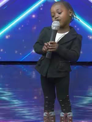 A young girl, dressed in a black blazer and leggings, sings into a microphone. She stands on a stage with a blue background lit with neon lights. The image captures a moment of performance.