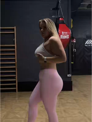 A woman in fitness attire is shown in profile within a gym setting. She wears pink leggings and a white sports bra, and her blonde hair is styled down. The gym's interiors features wooden floors, a boxing bag, and training equipment, creating a sporty vibe. Shadows and dim lighting provide depth, while the woman's stance suggests movement, possibly a dance.