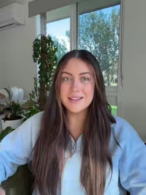 A woman with long, dark hair and a fair complexion smiles directly at the camera. She's wearing a light blue top in front of glass doors and a window with sunlight streaming through. Plants are visible in the background.
