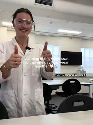 A young woman in a lab coat and safety glasses gives two thumbs up in a brightly lit lab setting with a sign in the background. There is text overlaid on the image.