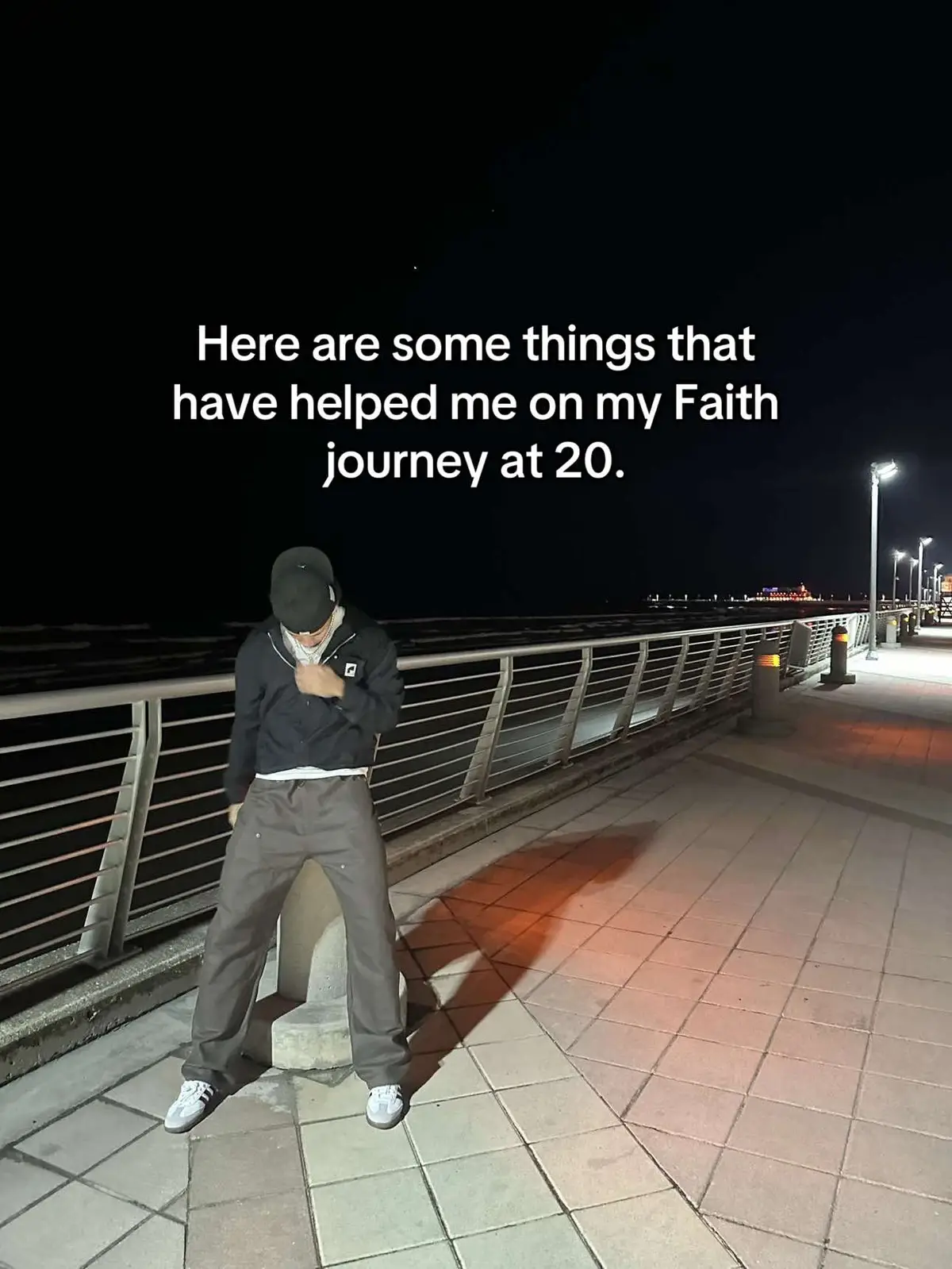 A nighttime shot of a young man standing on a boardwalk. He wears a black jacket, a black cap, and grey pants, with a white top showing under the jacket. The background is dark, but lights illuminate the pathway and the water edge. The text overlay reads: 'Here are some things that have helped me on my Faith journey at 20.'