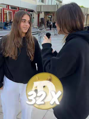 Two young women are standing outdoors, one speaking into a microphone toward the other. The woman on the right is holding the microphone. Both are wearing black sweatshirts and appearing to be in an outdoor setting. Overlaying the bottom is a yellow and gold circular graphic with a doge image, the letter D, and the number 32x.