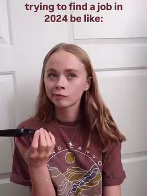 A young woman with blonde hair and a concerned expression holds a black object near her face, likely a phone or device. The background is a plain white wall. Text overlay reads 'trying to find a job in 2024 be like:'