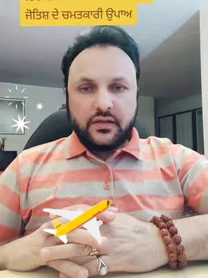 A man in an orange and white striped shirt holds a small toy airplane. He has a beard and is wearing a ring and a beaded bracelet. The background is blurred, but appears to be indoors. There is a yellow text box with text in a foreign language.