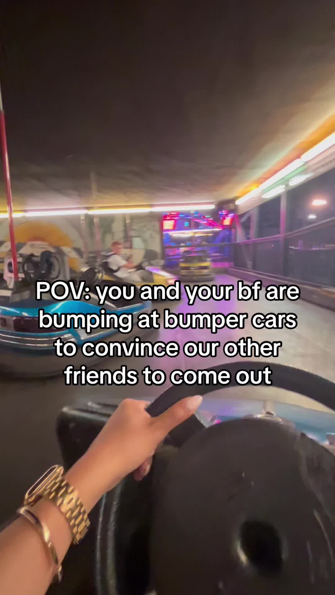 A first-person perspective shot inside a bumper car at an amusement park. The driver's hand, wearing a gold watch and bracelet, is visible on the steering wheel. The text overlay reads, 'POV: you and your bf are bumping at bumper cars to convince our other friends to come out.' The background shows the interior of what seem to be a bumper car arena, with other cars and people visible. The lighting is dim, and the overall mood seems fun and energetic.
