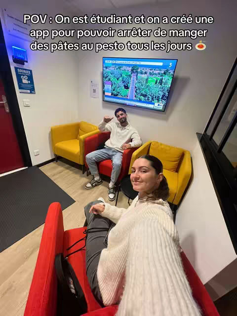 A selfie-style shot with two people in a room. A woman in a white sweater is in the foreground, seated in a red chair; her face is close to the camera. A man is pictured in the background, seated in a yellow chair, with another yellow chair behind him and a flat screen display on the wall. The background is a simple wall.