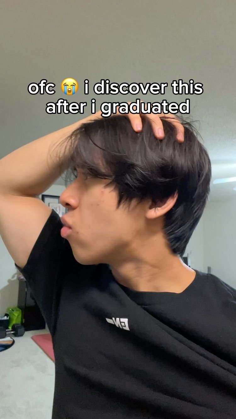 A young man with dark hair poses in a side profile, looking towards the camera with a slightly puckered expression, appearing contemplative. He wears a black t-shirt and has his hand up to his head, against a neutral indoor background.