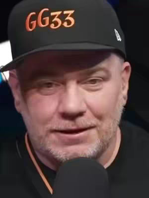 A man is looking at the camera wearing a black cap with the text "GG33" written on it in orange, his face is lit with average studio lighting, and he is holding a microphone.