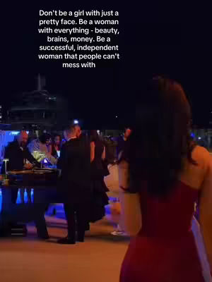 A woman with long black hair, viewed from the back and angled to the right side of the frame, is wearing a red dress and standing in front of a blurry outdoor gathering. Text overlays are present in the top portion of the image, and the background includes indistinct figures and lights.