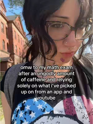 A selfie-style shot with a young woman in the foreground and a blurred background of a building. The woman is wearing large glasses, a black shirt with a stars and stripes design, and has a small tattoo near her mouth. The focus is on her face, and the text overlay provides context about a math exam.