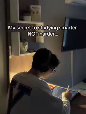 A man is seated at a desk, viewed from behind, with his back to the camera. He is wearing a white sweatshirt with dark accents on the back. He is wearing glasses and headphones. The lighting suggests a study or workspace with a display or screen visible on the right.
