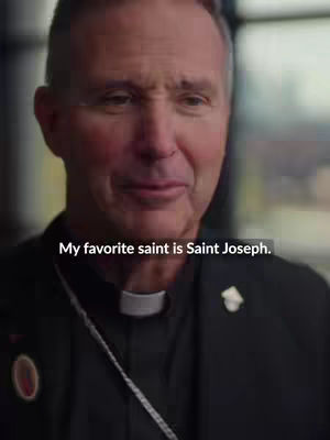 A close-up shot of a man wearing a black clerical collar and jacket. Text overlayed on image reads "My favorite saint is Saint Joseph."