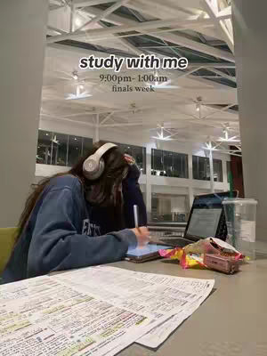 A woman is studying at a table with papers, a tablet, a laptop, and headphones. The background is a large open space with a high ceiling and bright lights.