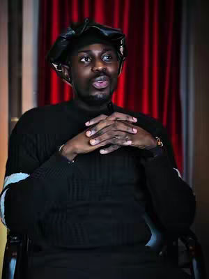 A close-up shot of a Black man gazing slightly upward, with hands clasped in front of his chest. He wears a black cap and a textured black sweater, set against a red curtain background. The lighting is soft, highlighting his features and the dark color palette.