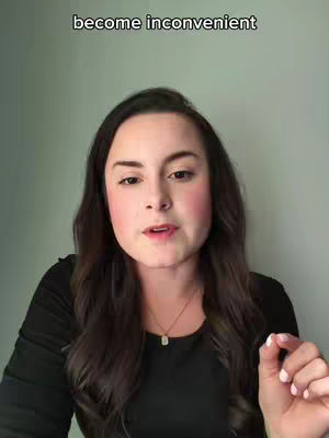 A woman in a black top is centered in the frame addressing the camera, with text overlayed at the top. She has dark hair, and is wearing a delicate necklace. Her skin has a pink flush on her cheeks, suggesting makeup. The background is plain, and the lighting is even.