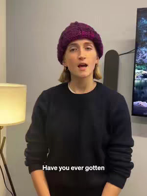 A woman wearing a purple knit hat and a black sweater is standing and talking. Text at the bottom reads 'Have you ever gotten'. A lamp is on the left and a monitor is on the right.