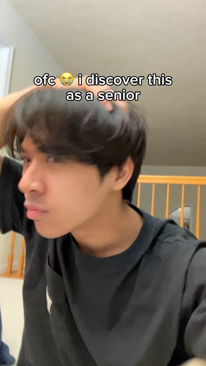 A young man with dark hair is shown in a medium shot, turned slightly to the side, looking towards the camera. His left hand is raised, touching his hair. The background is blurred, revealing a neutral, domestic interior with a staircase in the distance. The man wears a black t-shirt, and text overlays on the image.