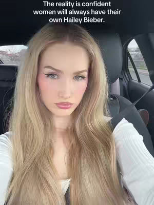 A woman with long blonde hair is seated in a car. The image is a close-up shot, focusing on her face and upper body. She appears to be looking at the camera with a neutral expression. The background shows the interior of the car and a glimpse of an outdoor scene through the window. The text overlay is present at the top.