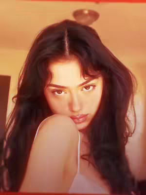 A young woman with long, wavy black hair gazes directly at the camera with a soft, inviting expression. She wears a white tank top and has a delicate hand near her shoulder. The lighting is warm and golden, adding to the intimate feel of the image. The background is blurred, emphasizing the woman's face.