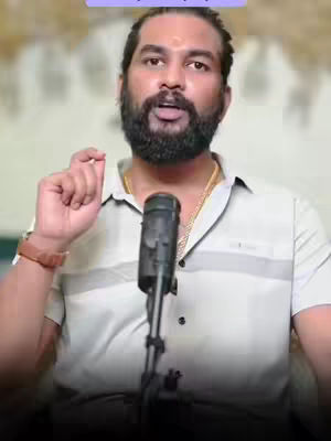 A man with a beard is holding a microphone towards his mouth, with one hand gesturing upwards in front of a blurred background of a light beige color. He is wearing a white collared shirt and a gold chain, with a brown watch on his wrist