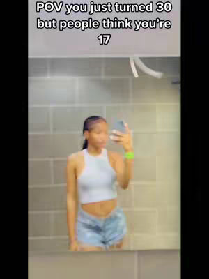 A woman takes a selfie in a bathroom mirror. The mirror reflects a woman in a white tank top and denim shorts. The background is a tiled wall. The image has text overlayed at the top of the screen.