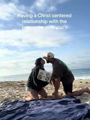 Two people are kissing on a beach in front of the ocean. The sky is cloudy and a blanket is in the foreground. The person on the left is holding a sign with handprints on it.