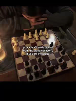 The image shows a chess board with chess pieces set up for a game. A person is leaning over the board, their head and upper body are visible, but their face is not clearly seen. The lighting appears dim, creating a somewhat moody atmosphere. The text overlay says: "pov you are so detached from the world you worry if you are even here".
