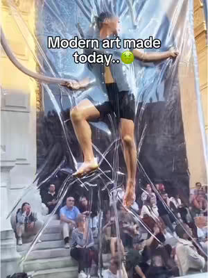 A man stands suspended within a large, transparent plastic structure. The man is wearing casual attire and is positioned as if he is climbing or hanging from the interior of the plastic. Below, a crowd of people watches, adding scale to the unusual modern art installation.