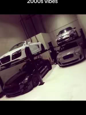 The image features several luxury cars parked in a garage setting. The cars are on display, and the image has a vintage, slightly desaturated feel, like something from the early 2000s. The composition suggests a focus on wealth and luxury.
