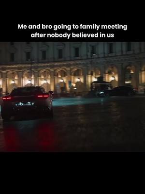 A nighttime shot of two cars driving away from the camera on a wet city street. The lead car has its brake lights on, and the background shows a large building with arched windows and lights.