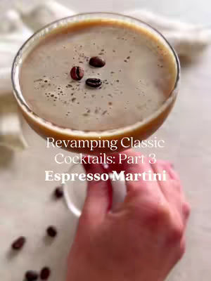 The image shows a close-up view of a non-alcoholic espresso martini in a stemmed glass. The drink is a light brown color and topped with a thin layer of foam. Three coffee beans are placed on top. The drink is held by a person's hand, with the background a blurred light color. The text overlayed on the image says 'Revamping Classic Cocktails: Part 3 Espresso Martini'.