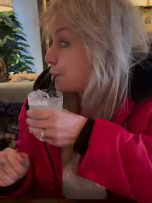 A woman with blonde hair is side-profile in an interior setting, drinking from a glass with a straw. There's ice in the drink, and she's wearing a red jacket. The background shows a glimpse of a decorated space.