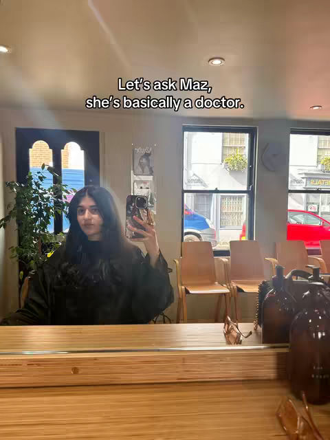 A mirror selfie taken in what appears to be a hair salon. The mirror reflects the room, with a wooden table in front of the mirror. The creator, a woman with dark hair, is holding her phone up to take the photo. The background shows chairs and windows with outside views.