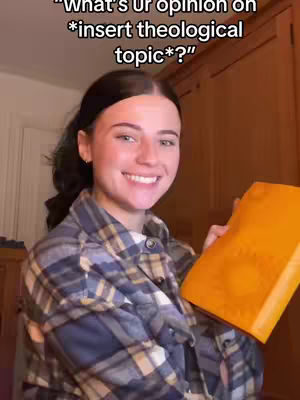 A young woman with dark hair and a plaid shirt is smiling, holding an orange book. The background includes a wooden cabinet and wall.