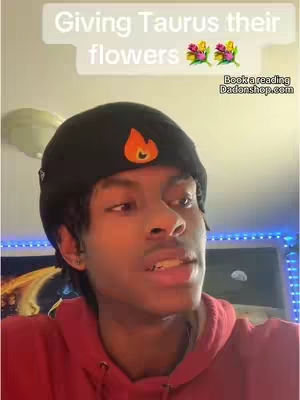 A young man with dark skin and a black beanie with a flame logo is talking to the camera. Above him is a text bubble with the words "Giving Taurus their flowers" and some flower emojis. The background shows a blurred-out room.
