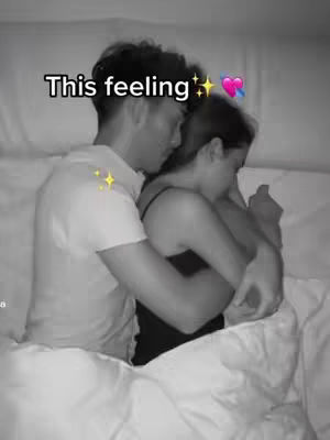 A black and white photo of a couple cuddling in bed. The man is on the left wearing a white shirt, hugging the woman, who is partially visible on the right and wearing a black tank top. They are under a white duvet. Above the couple, the text overlay "This feeling" is displayed.
