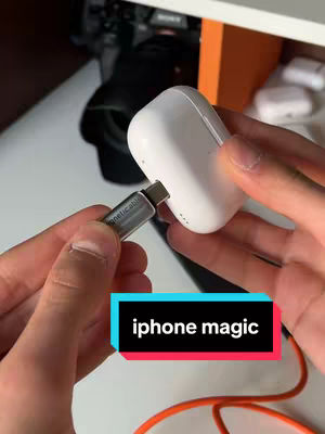 A person's hands are holding a white AirPods case and plugging a USB-C cable into it. The AirPods case is positioned in the center of the frame, with the cable on the left. The background includes a white desk, an orange cable, and objects out of focus. A text overlay says 'iphone magic'.