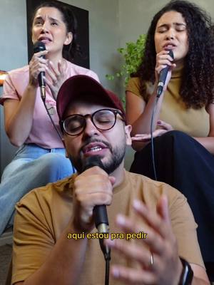A group of three people are singing into microphones. In the foreground is a man wearing glasses and a cap, framed from the chest up, singing with his eyes closed while holding a microphone. Behind him, two women are also holding microphones and singing. The background is a soft, warm-toned space, suggesting an indoor setting, and has a plant to the right.