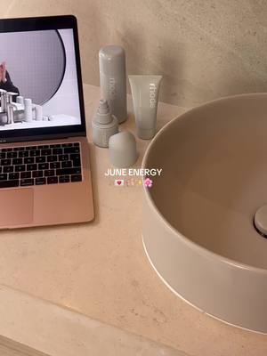 The image presents a bathroom counter setting with elements suggesting a self-care or beauty-related theme. A laptop, open and displaying a screen, sits on the left. Beside the laptop are several skincare products and a sink to the right.