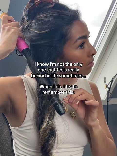 A woman with long, dark hair is featured, curling her hair with a pink curling iron. The background is a light-colored wall, and text overlays appear in the frame.