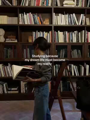 A well-lit image depicts a woman in a library. She is standing in front of a large dark wooden bookshelf filled with books. She is reading from a large book, the pages wide open. The text overlay states "Studying because my dream life must become my reality"