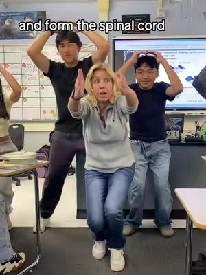 The image captures a classroom scene featuring three individuals seemingly acting out a biology concept. A woman in the center is the focal point, flanked by two men on either side. They are indoors, with a whiteboard and a large screen visible in the background.